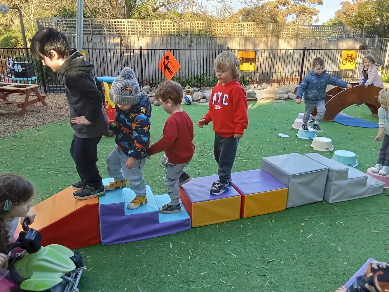 Children at a childcare centre engage in outdoor play, balancing on a colourful soft foam obstacle course set up on artificial grass.
