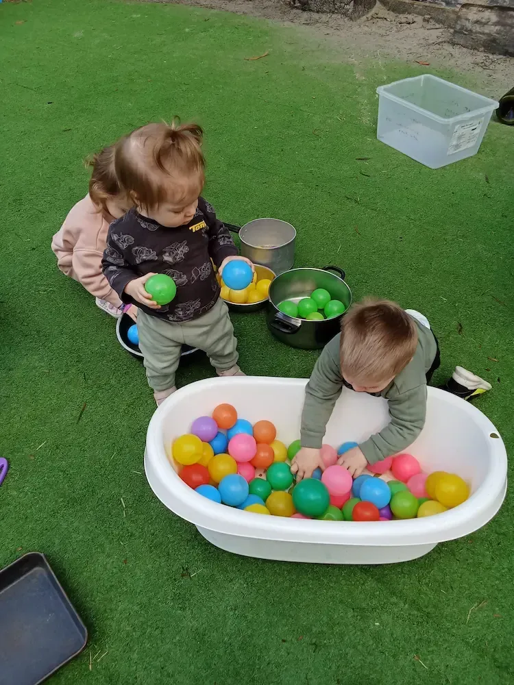 Toddlers at a childcare center engage in sensory play outdoors, exploring a plastic tub filled with colorful plastic balls.