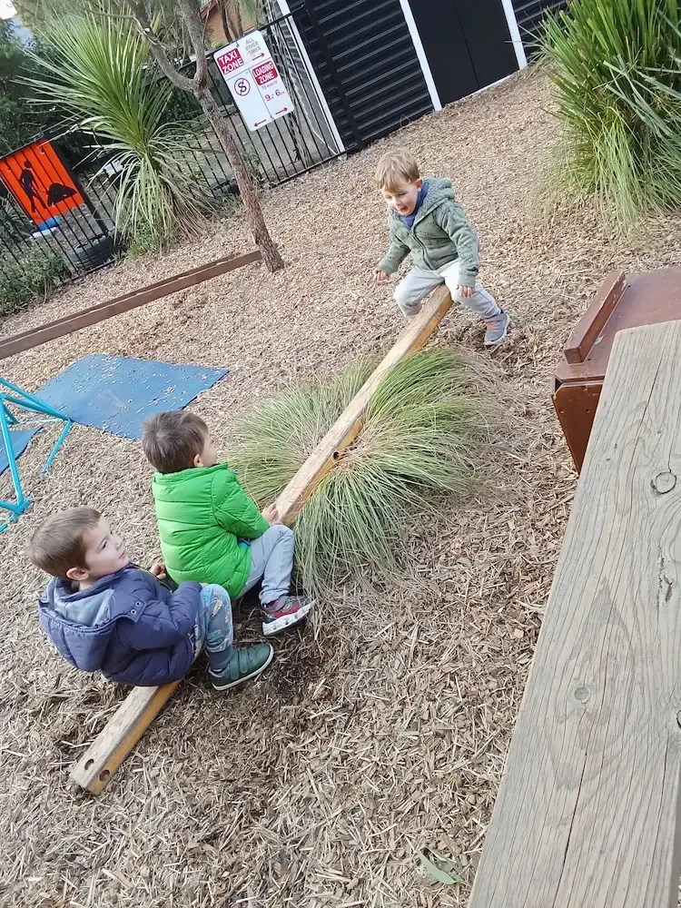 Three young children at a childcare center play outdoors on a natural wooden seesaw surrounded by bark mulch and native plants. 