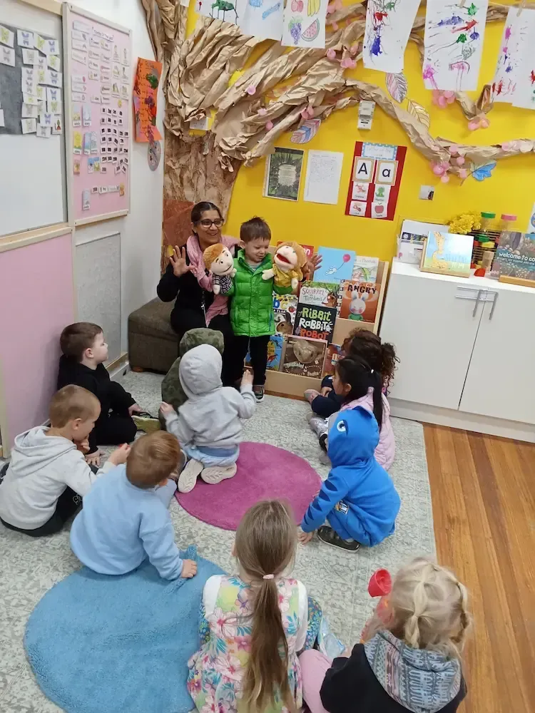 A childcare classroom scene showing a group of young children sitting on colourful mats, attentively watching a boy in a green jacket holding two hand puppets.