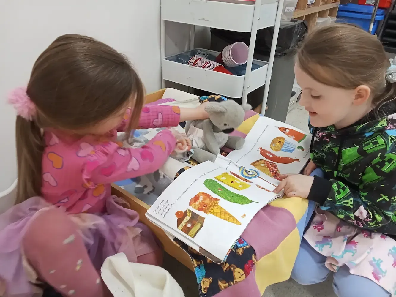 Two young girls at a childcare center sit together reading a colorful picture book, with one holding a stuffed animal. 
