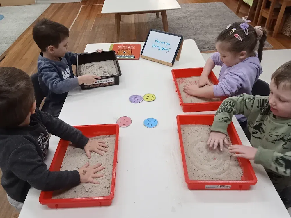 Young children at a childcare centre engage in a sensory activity, using their hands to explore trays filled with sand.