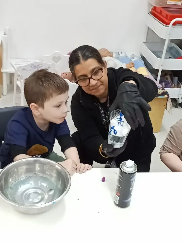 A childcare educator demonstrates a science experiment to a curious young boy, using a clear plastic cup filled with water, shaving cream, and blue dye.