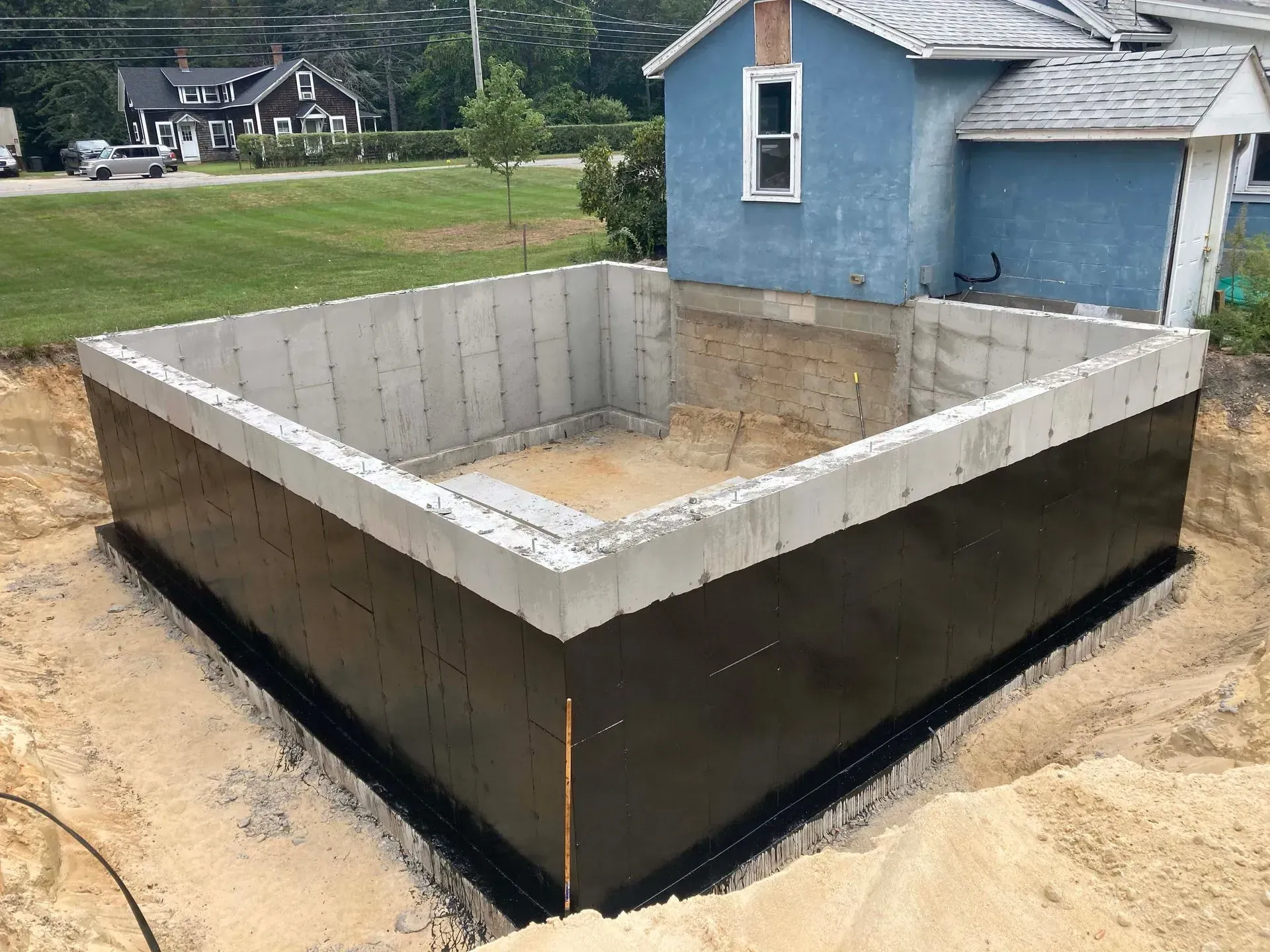 Foundation under construction beside blue house, with concrete walls and excavated dirt around it