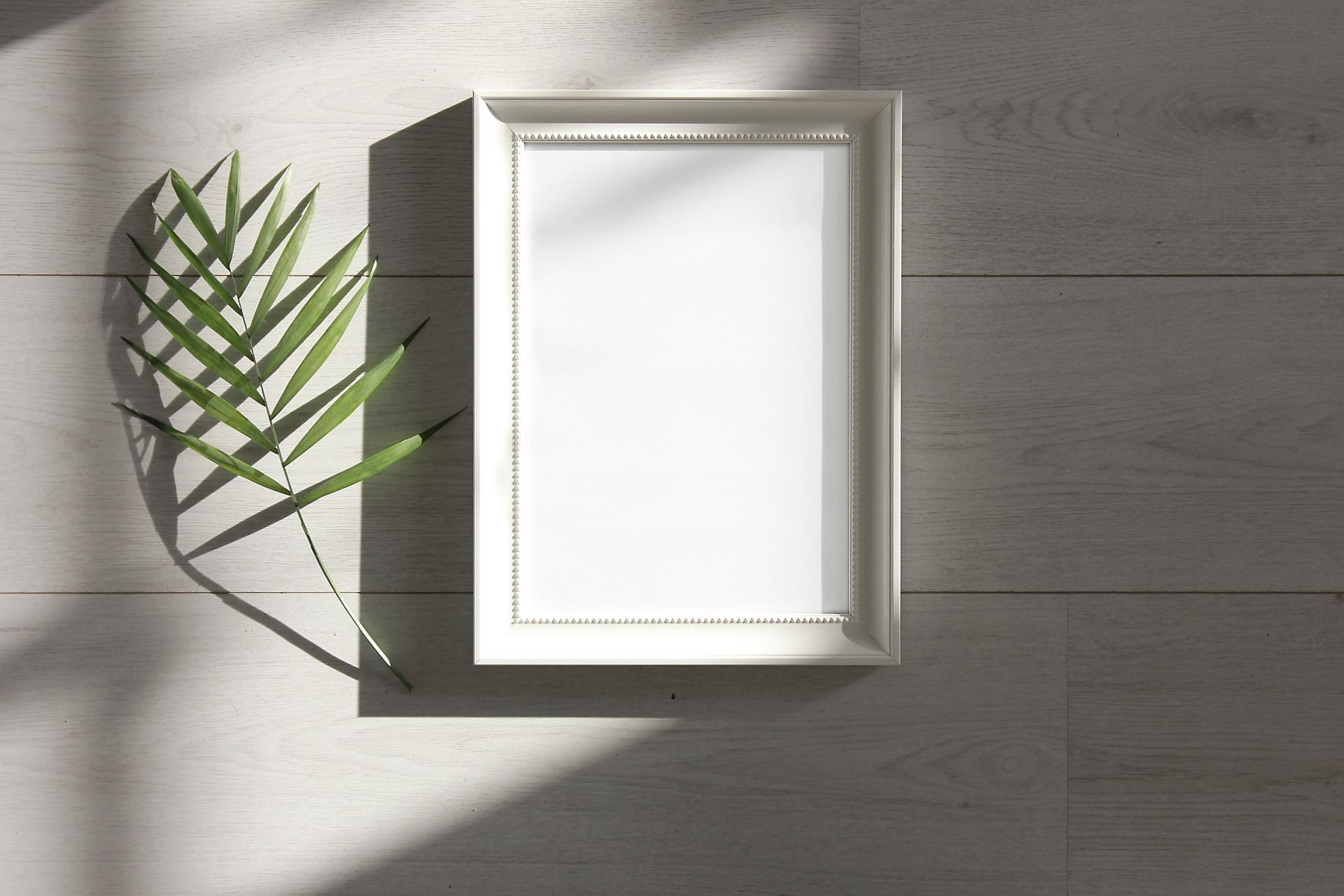 A white picture frame is sitting on a wooden floor next to a palm leaf.