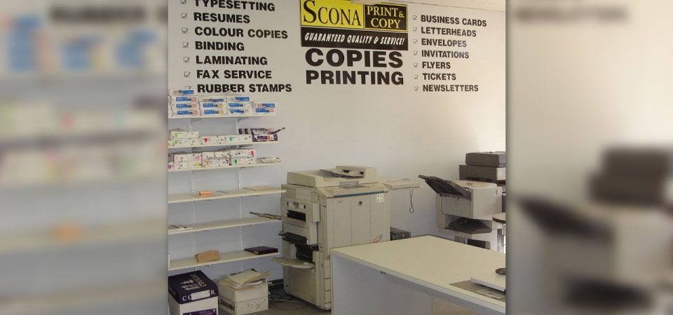 A room with a printer , desk , shelves and a sign on the wall that says copies printing.