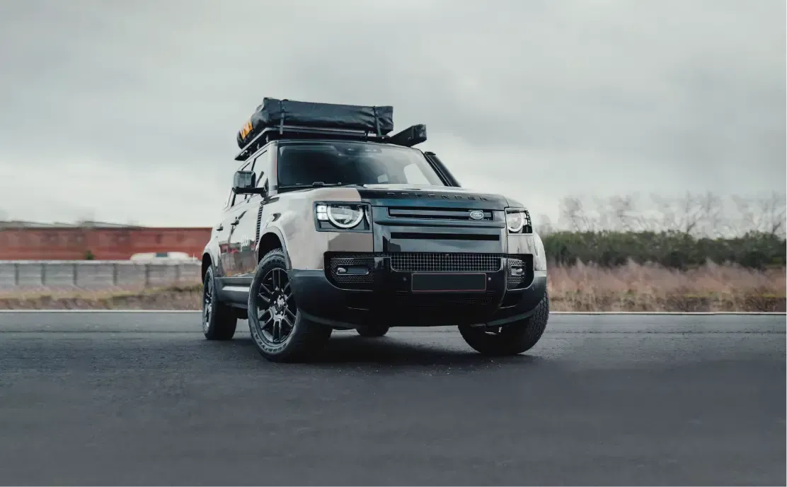 A land rover defender with a tent on top of it is driving down a road | Redline Automotive