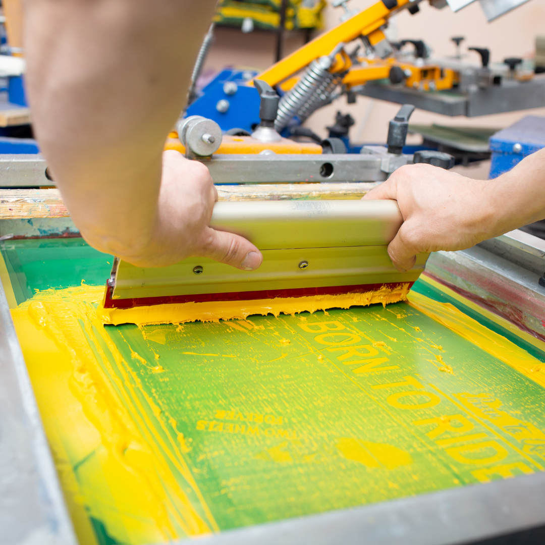 Hands pressing a squeegee across a silkscreen, spreading yellow ink.