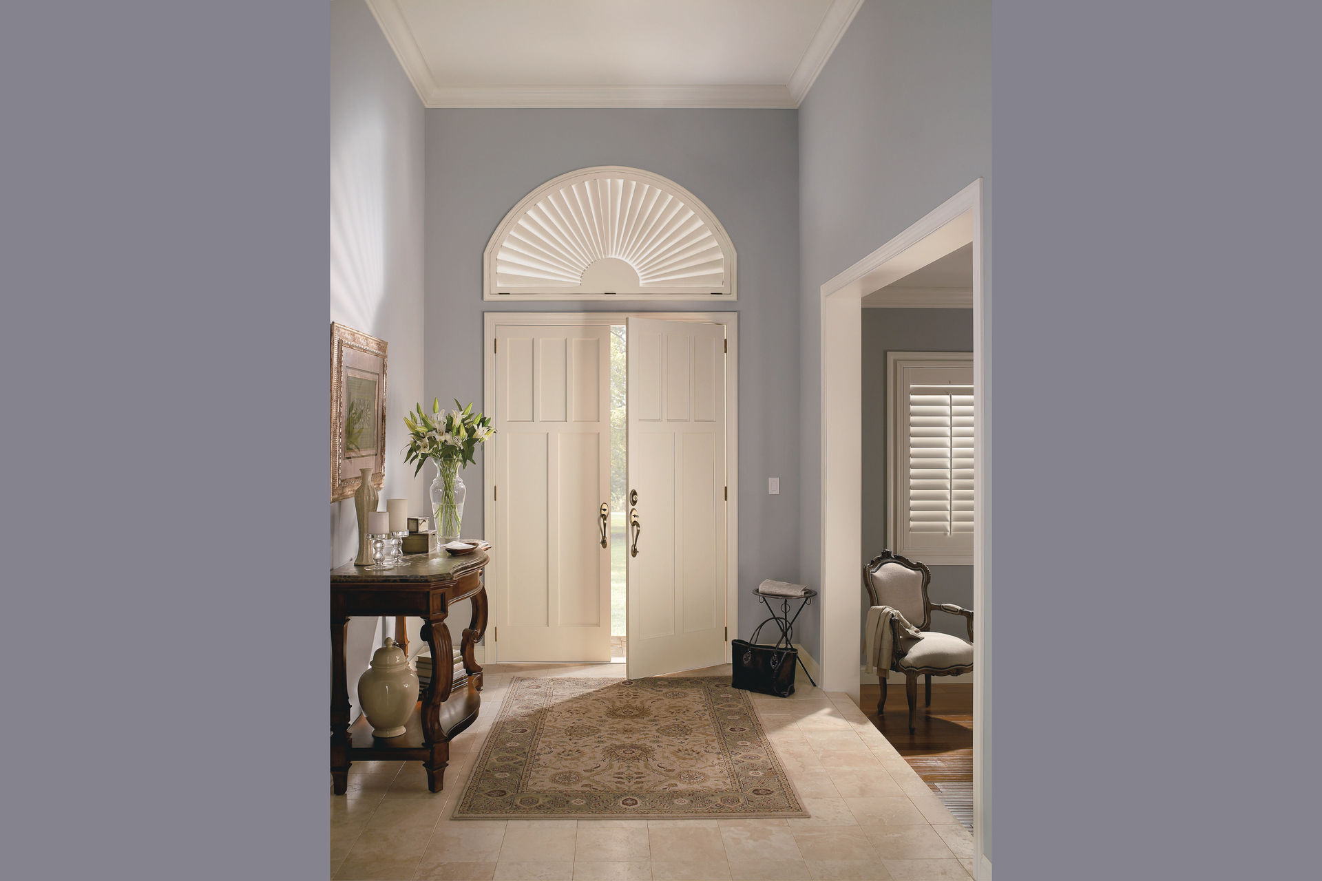 A sunlit entryway features double white doors with a fanlight, a wooden console table, and a rug on stone flooring.