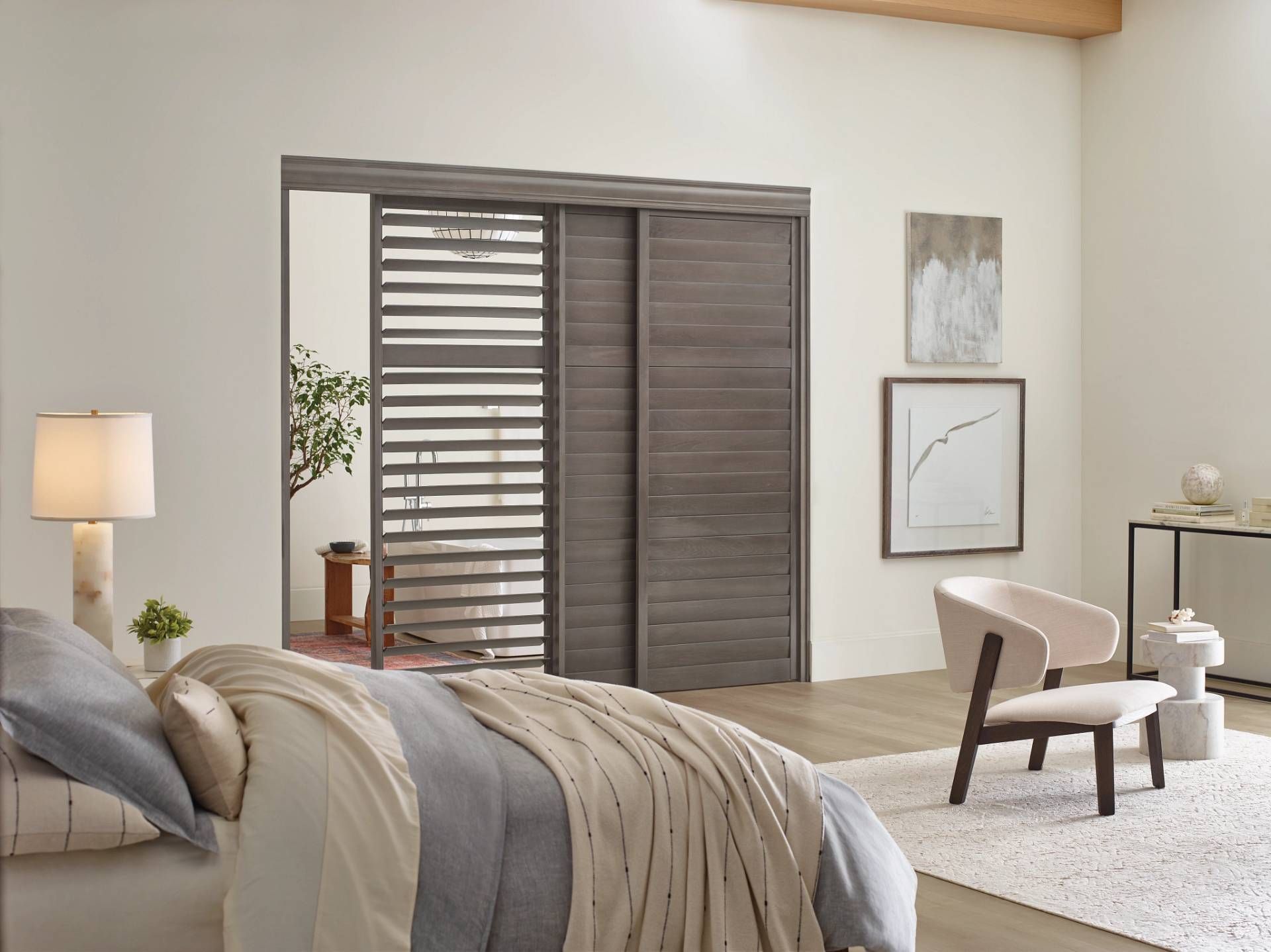 A bright, modern bedroom featuring a sliding, slatted dark wood door, a bed with neutral linens, and a cream accent chair.