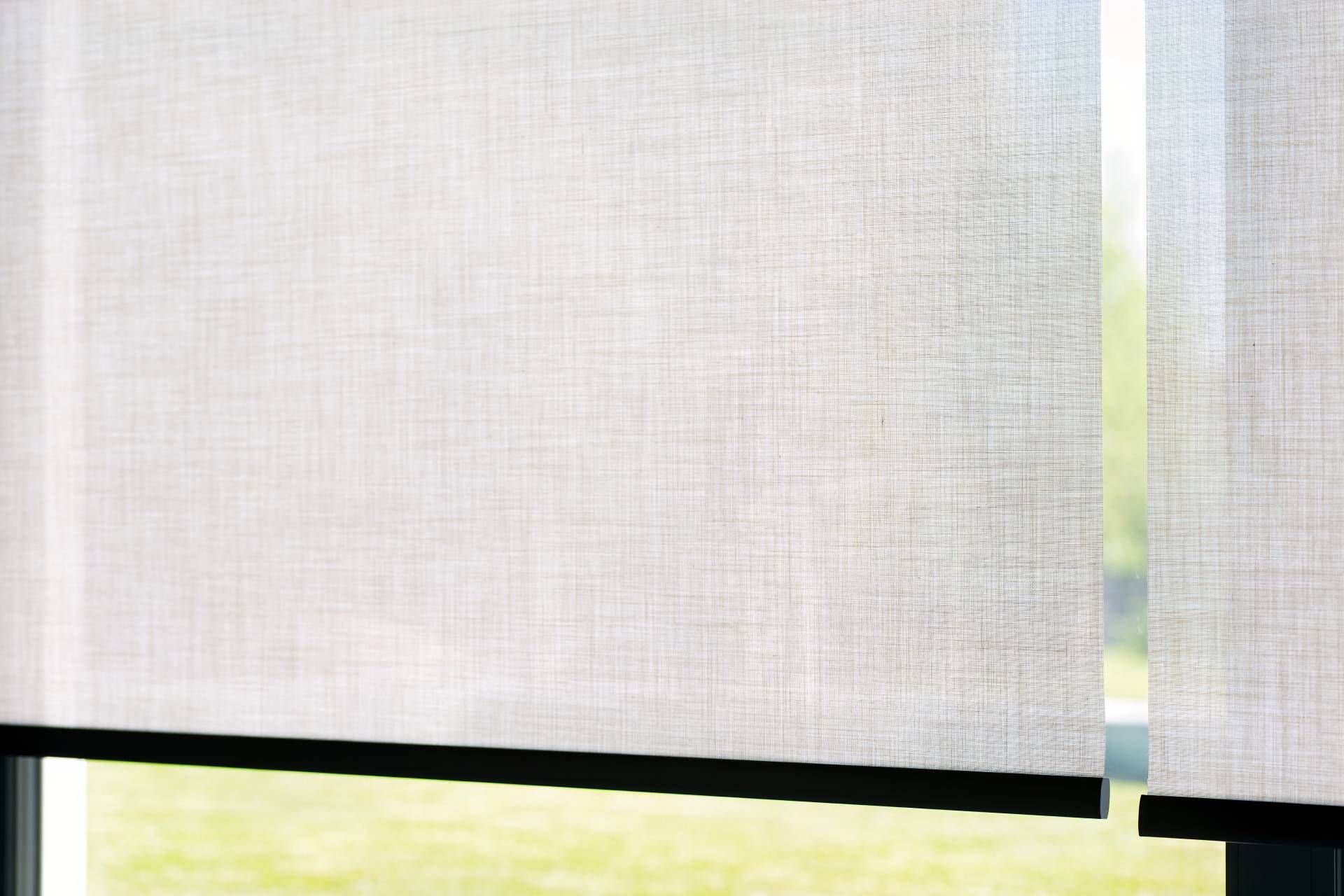 Two light-colored, textured roller shades hang in front of a window, partially revealing a green outdoor landscape.
