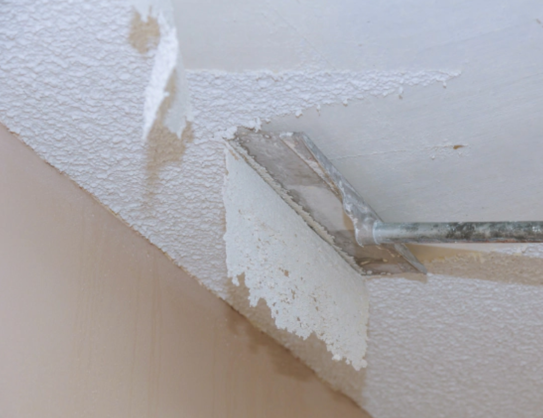 Scraper removing peeling white paint from a ceiling corner.
