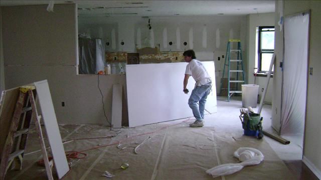Man installing drywall in a room under construction, standing on a floor covered with drop cloths.