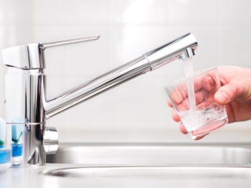 A person filling a glass with water from a shiny chrome faucet in a kitchen sink.