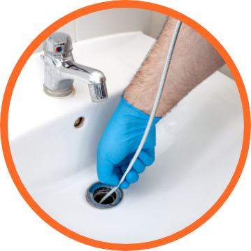 A person wearing a blue glove unclogs a bathroom sink drain with a plumbing snake.