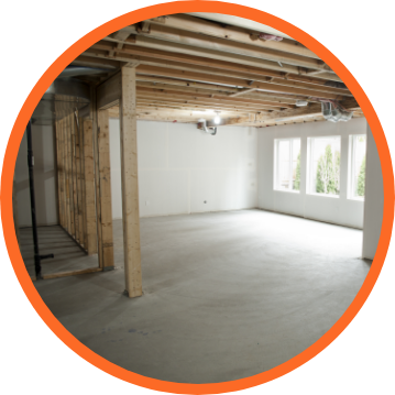 Unfinished basement with concrete floor, wooden beams, and framed walls. Bright windows on the right.