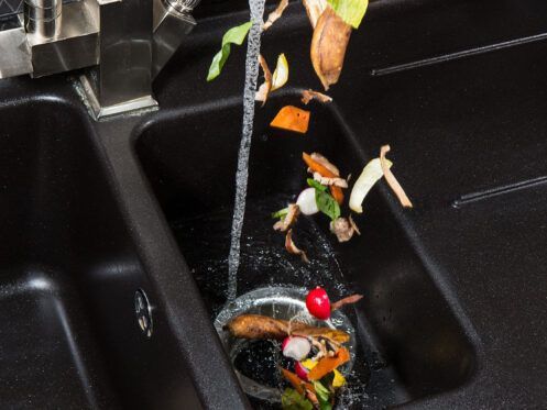 Food scraps and water pouring into a garbage disposal in a black kitchen sink.