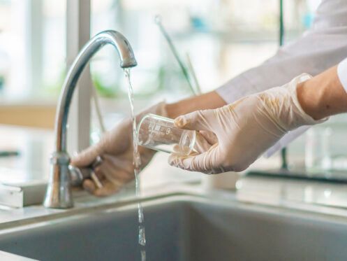 Person in lab coat and gloves filling a beaker with water from a faucet in a lab sink.
