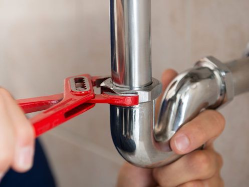 A person's hands using a red adjustable wrench to tighten a chrome-colored pipe in a bathroom setting.