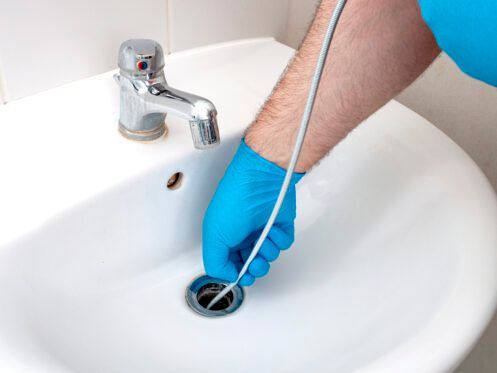 Person in blue glove using a drain snake to unclog a white bathroom sink.