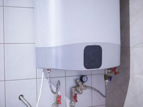 White electric water heater mounted on a tiled wall, with pipes and valves visible.