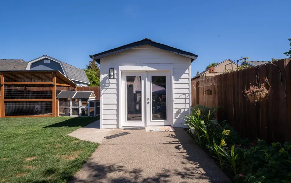 A small white house is sitting in the backyard next to a wooden fence.