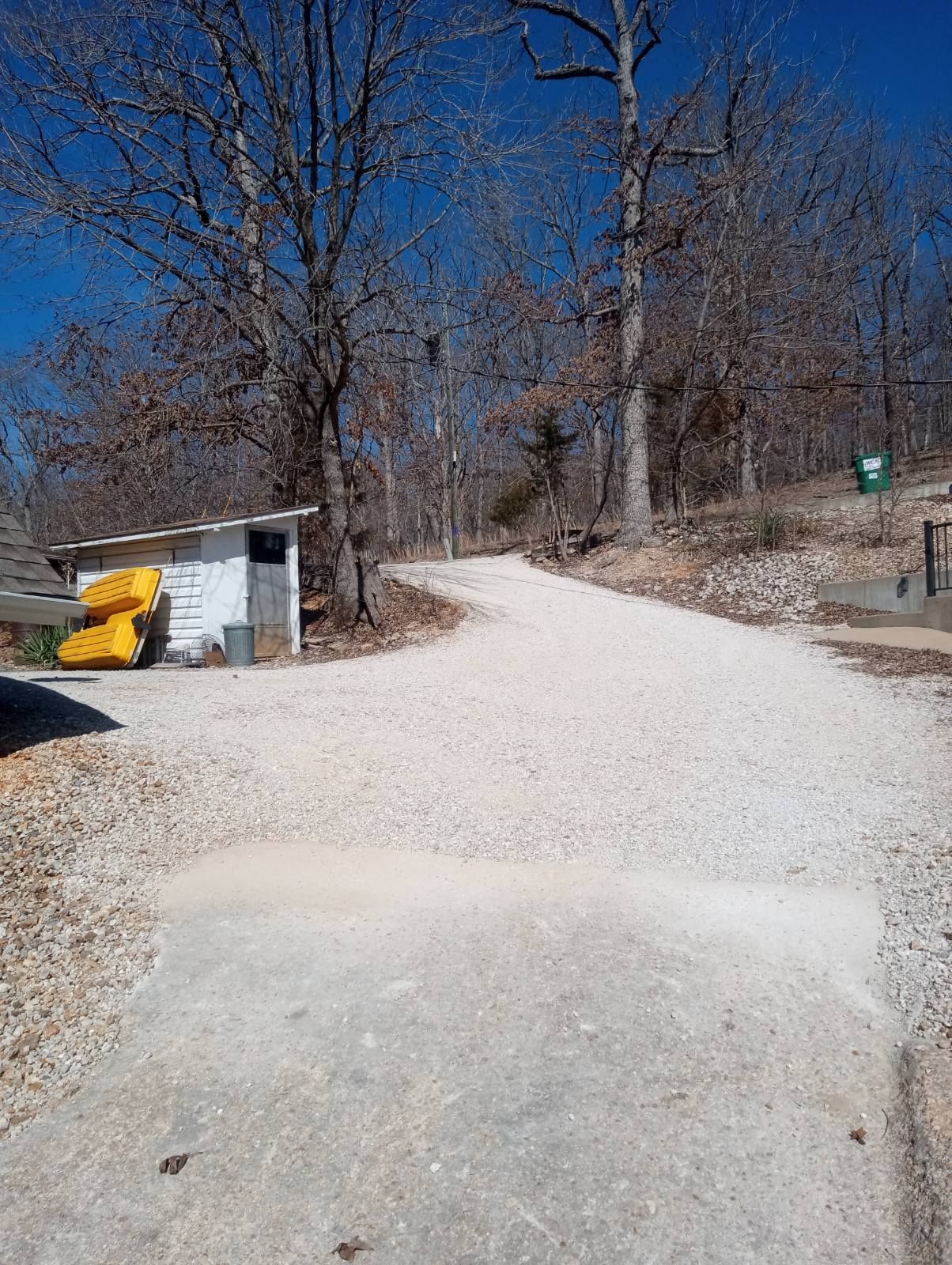 A gravel road curves through a wooded area, passing a small white building and trees under a blue sky.
