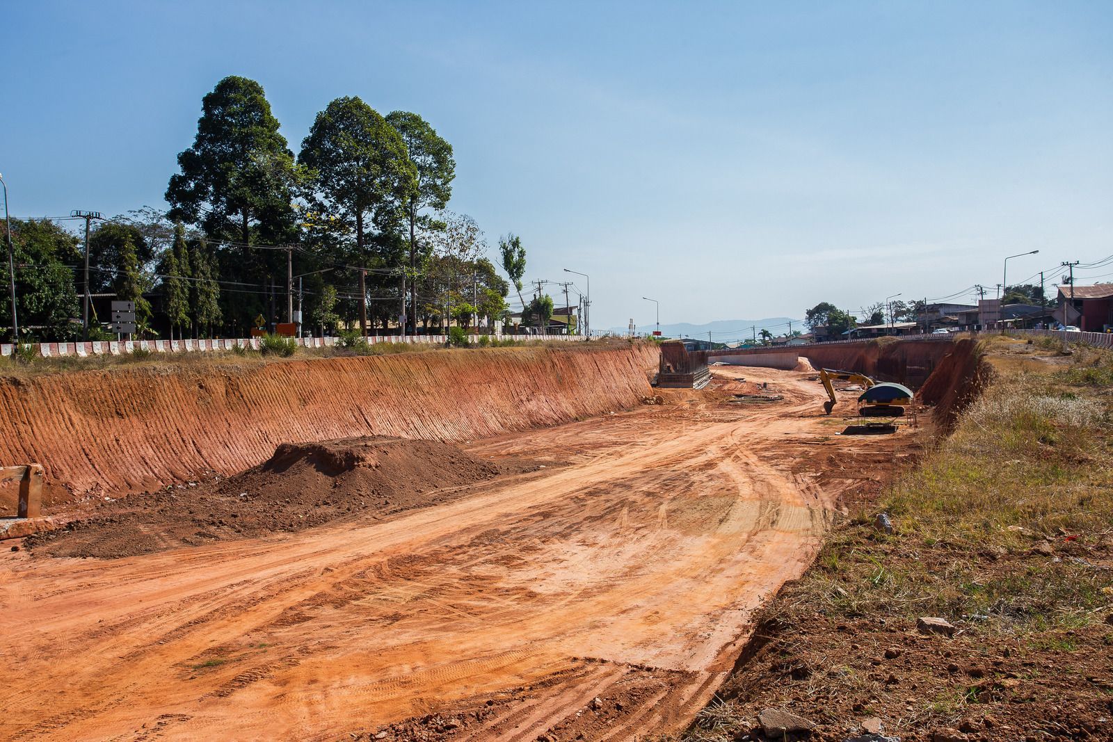 Construction site with deep red-brown dirt, road being built, blue sky, trees in the background.