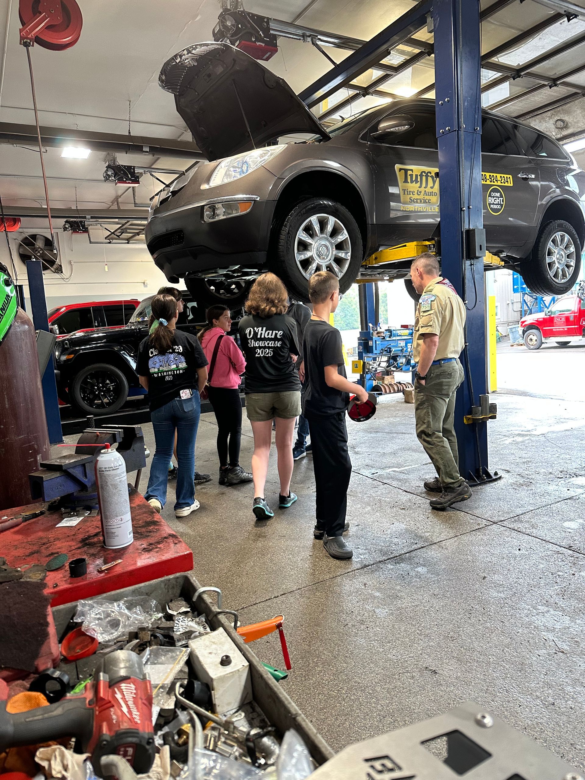 Group of people in a garage, inspecting a car on a lift; a mechanic speaks to them.
