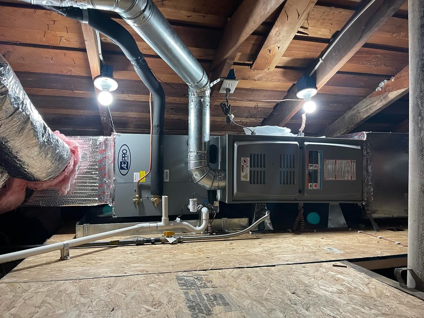 HVAC unit installed in an attic; metallic ductwork and pipes are visible with exposed wooden beams.