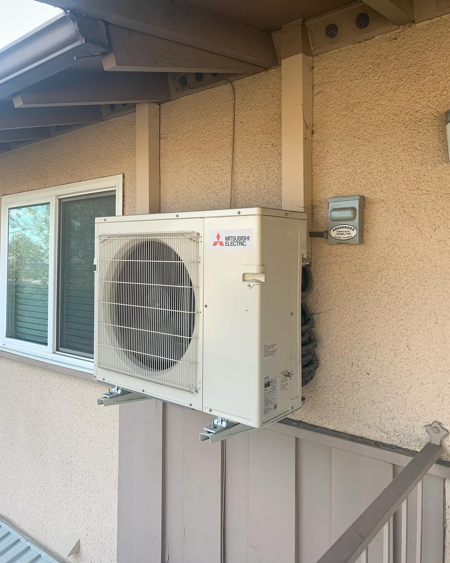 Exterior view of an air conditioning unit attached to a stucco wall next to a window and electrical meter.