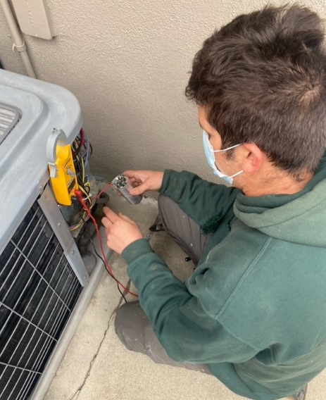 Person in a green hoodie repairs an AC unit, using a multimeter. Outdoors, near a beige wall.