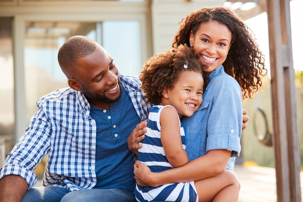 Family hugging outdoors, smiling. Man in blue shirt, woman in denim, child in striped dress.