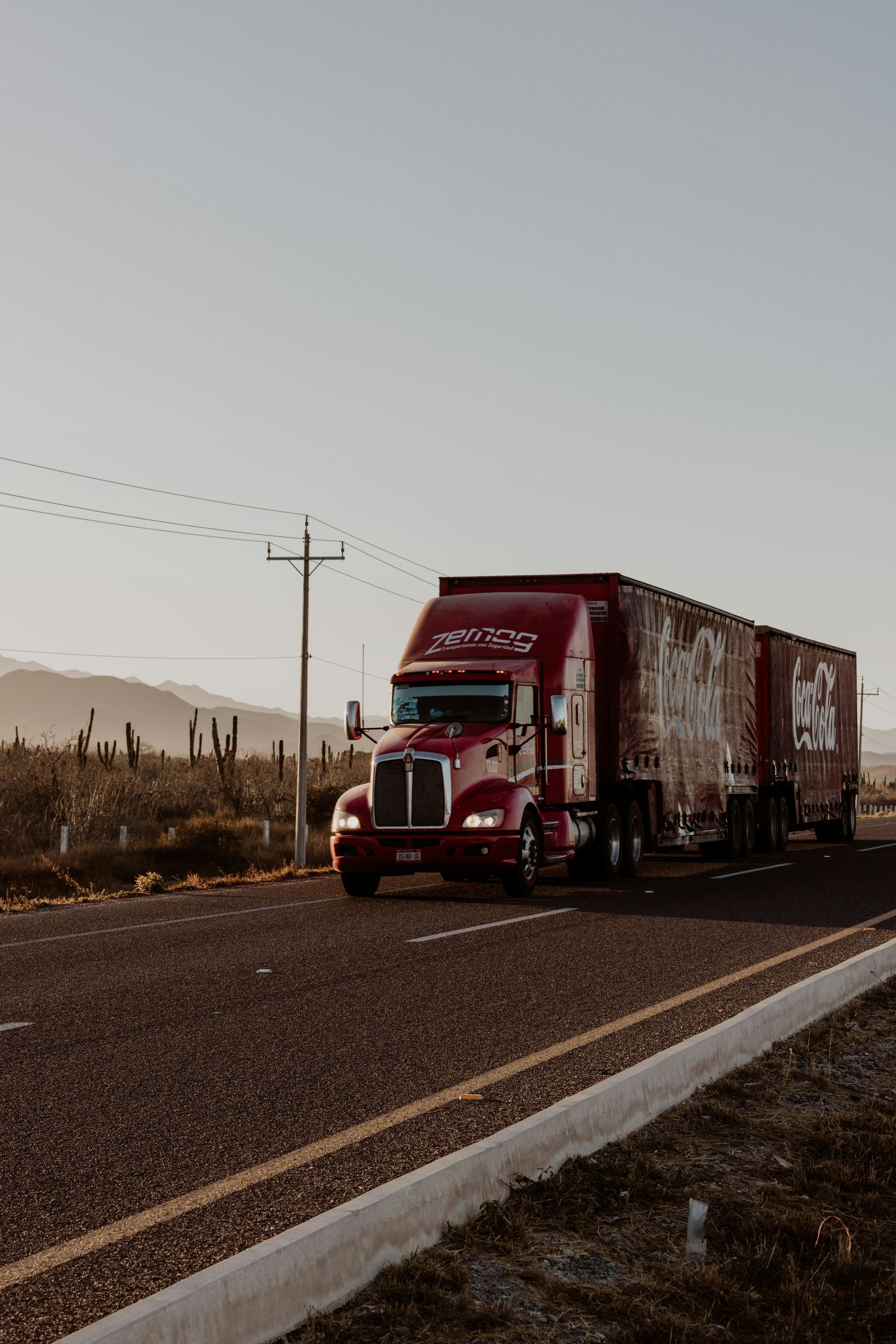 Red Coca-Cola semi-truck driving on a desert highway under a clear sky.
