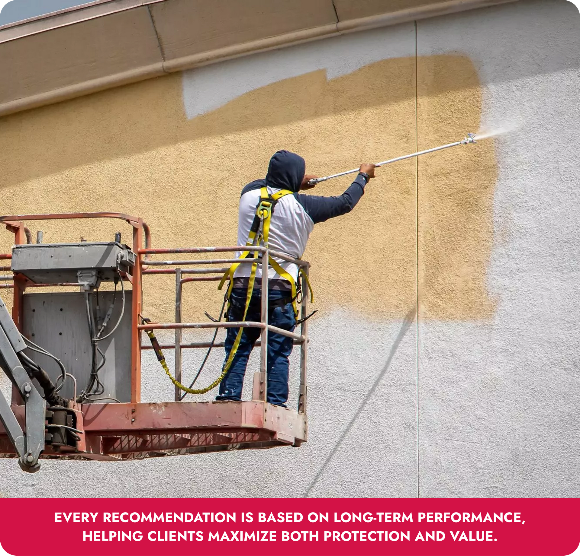 Worker on a lift spraying beige coating onto an exterior wall near a gray section
