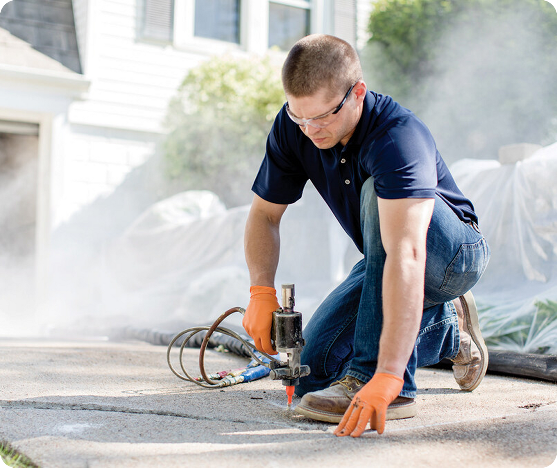 Worker in orange gloves uses a power saw to cut concrete on a driveway, with dust rising around him.