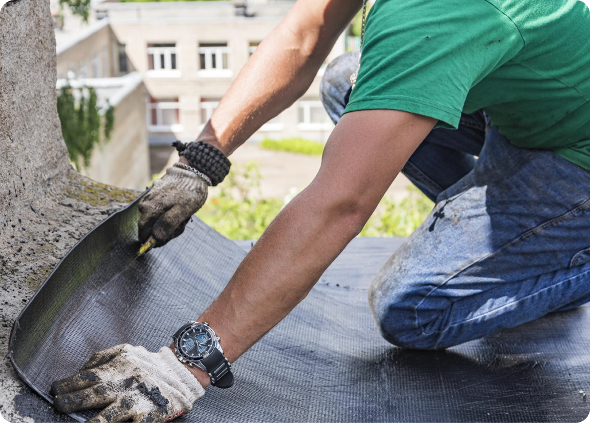 Worker kneeling on a rooftop, smoothing black roofing membrane with gloved hands.