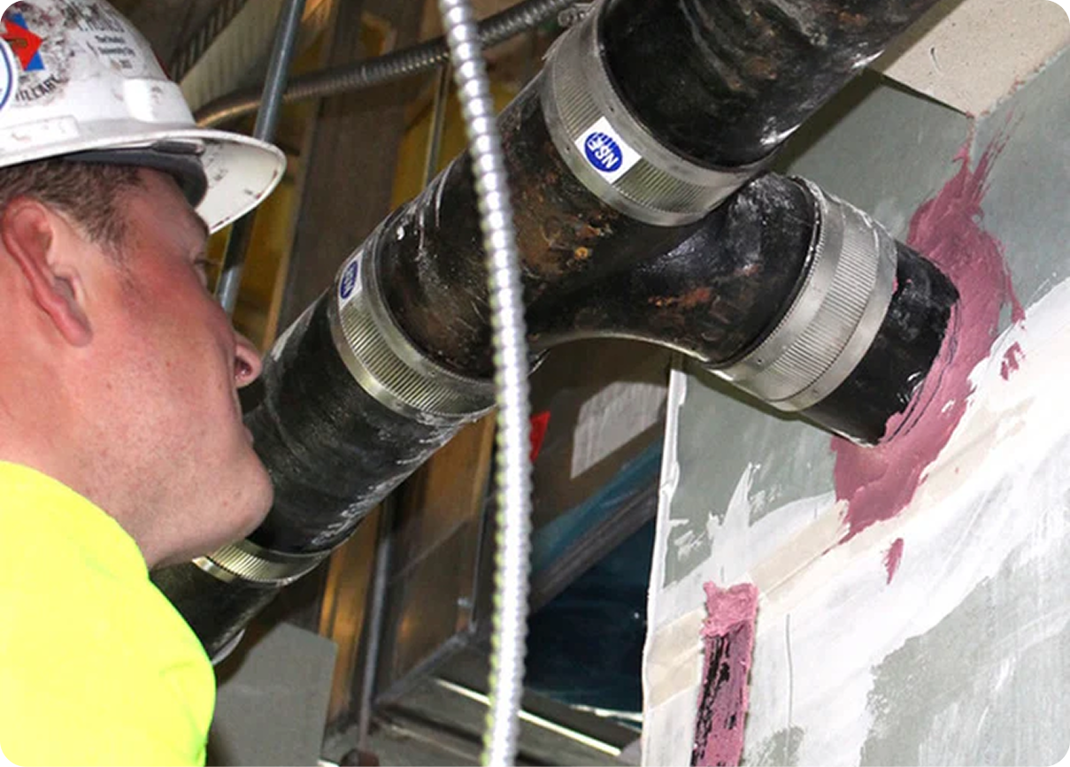 Worker in a hard hat inspecting large black duct pipes in a ceiling space