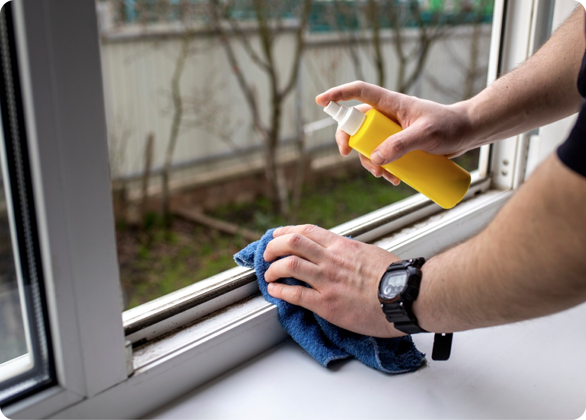 Hands cleaning a window sill with a blue cloth and yellow spray bottle