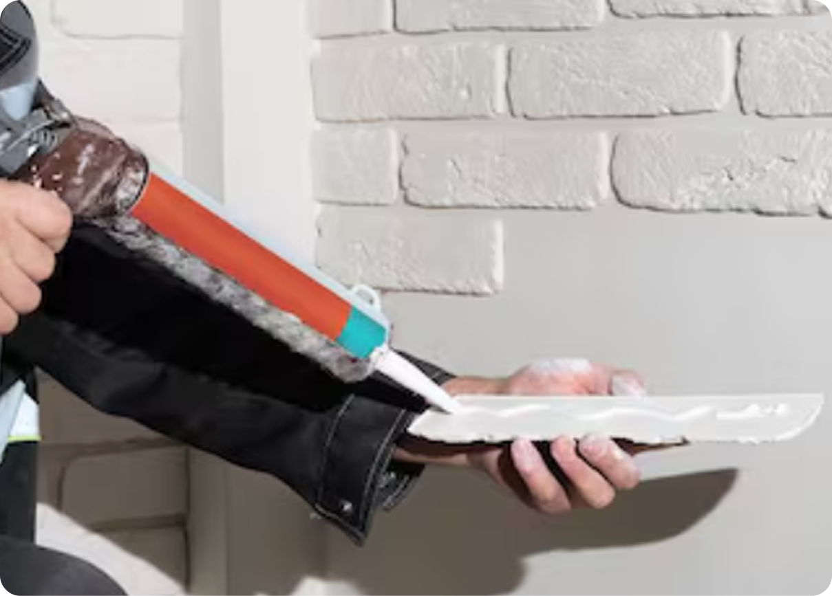 Hand applying caulk with a red caulking gun to a white tray against a white brick wall