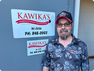 Man standing beside a Kawika’s sign on a gray wall, wearing a cap and glasses, smiling slightly.