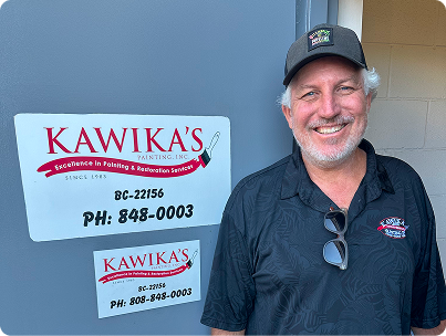 Man smiling beside Kawika’s plumbing sign on a blue wall, wearing a black cap and polo shirt