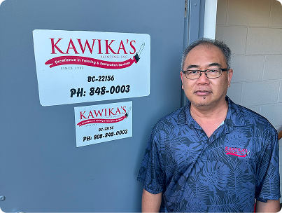 Man standing beside a KAWIKA’S sign on a gray wall, wearing a blue patterned shirt.