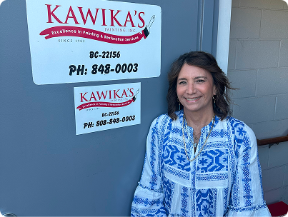Woman standing beside a Kawika’s sign on a blue wall, wearing a blue patterned top and smiling.
