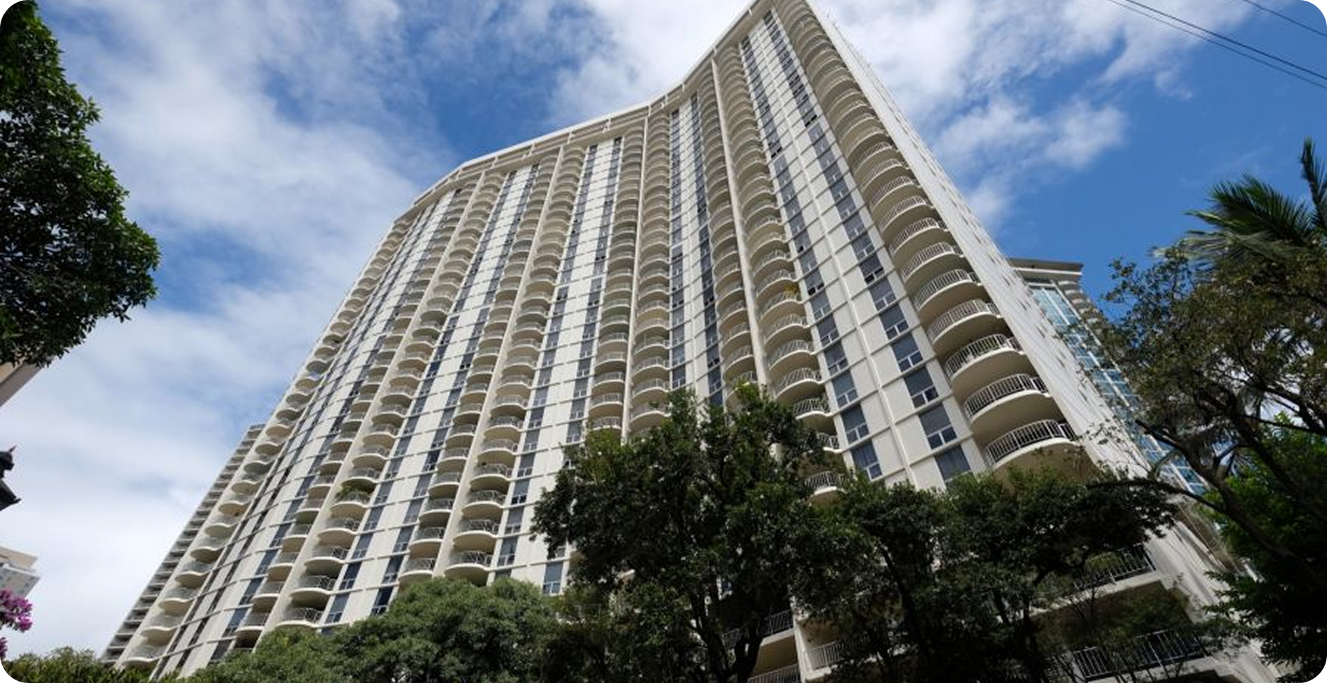 Tall beige high-rise apartment building viewed from below, framed by trees against a cloudy blue sky