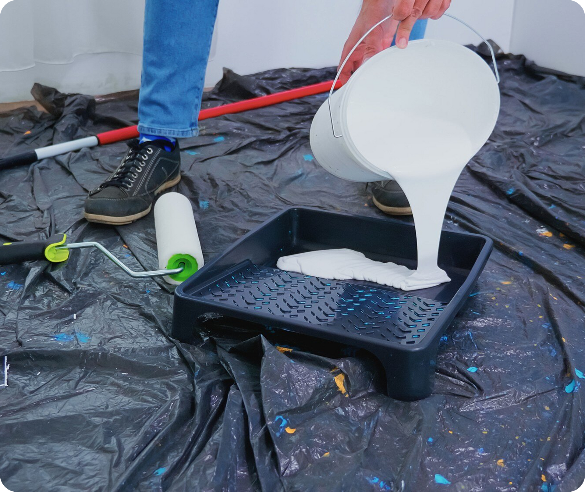 White paint pours from a bucket into a black tray on a tarp-covered floor.