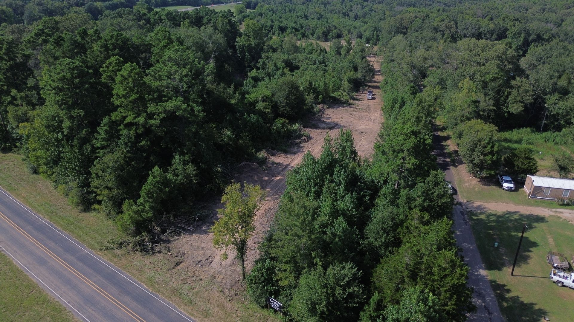 Aerial view of a road bordered by trees; cleared land and structures are visible.