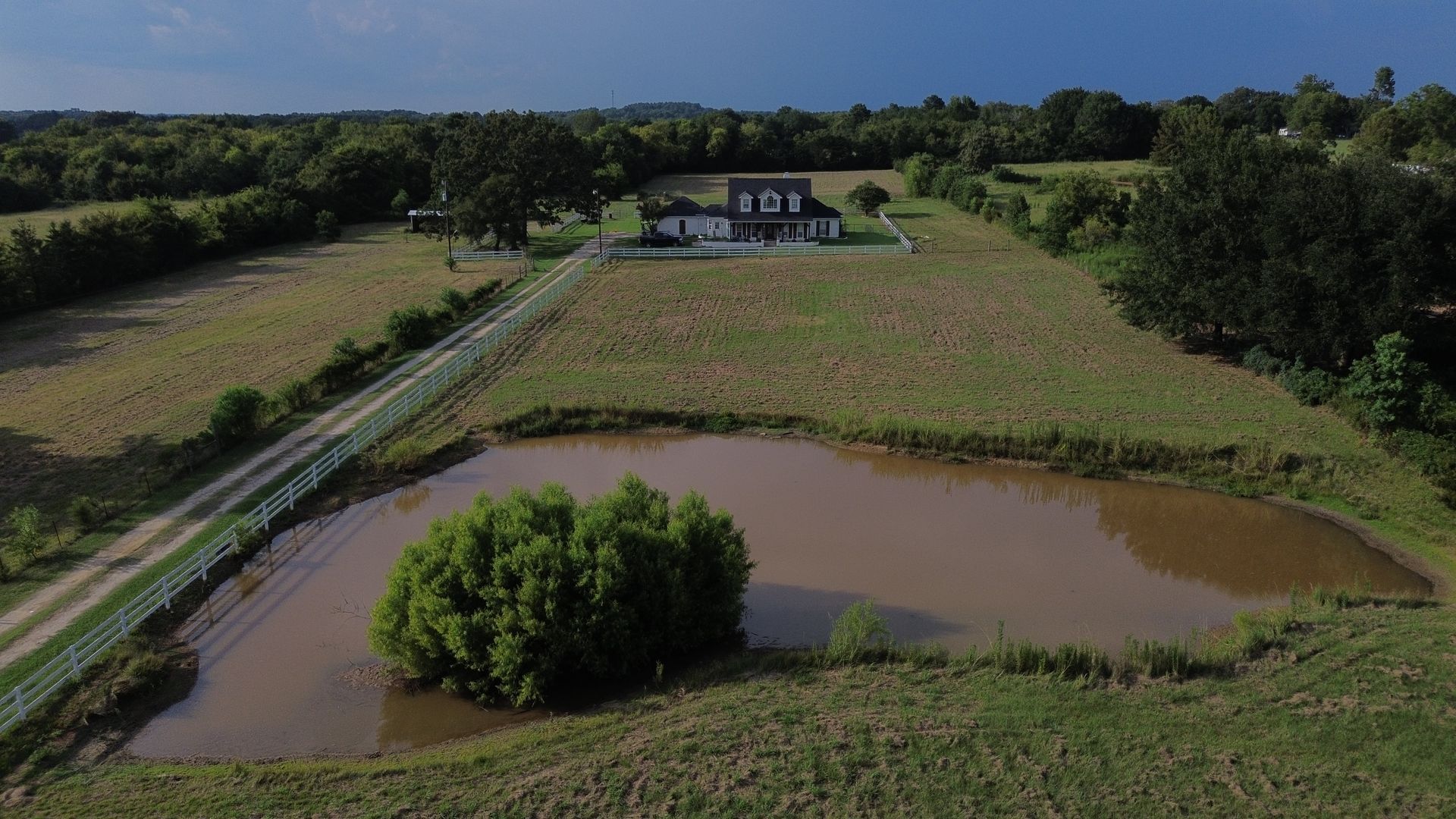 Elevated view of a house on a grassy hill, a long road leading to it, and a pond in the foreground.