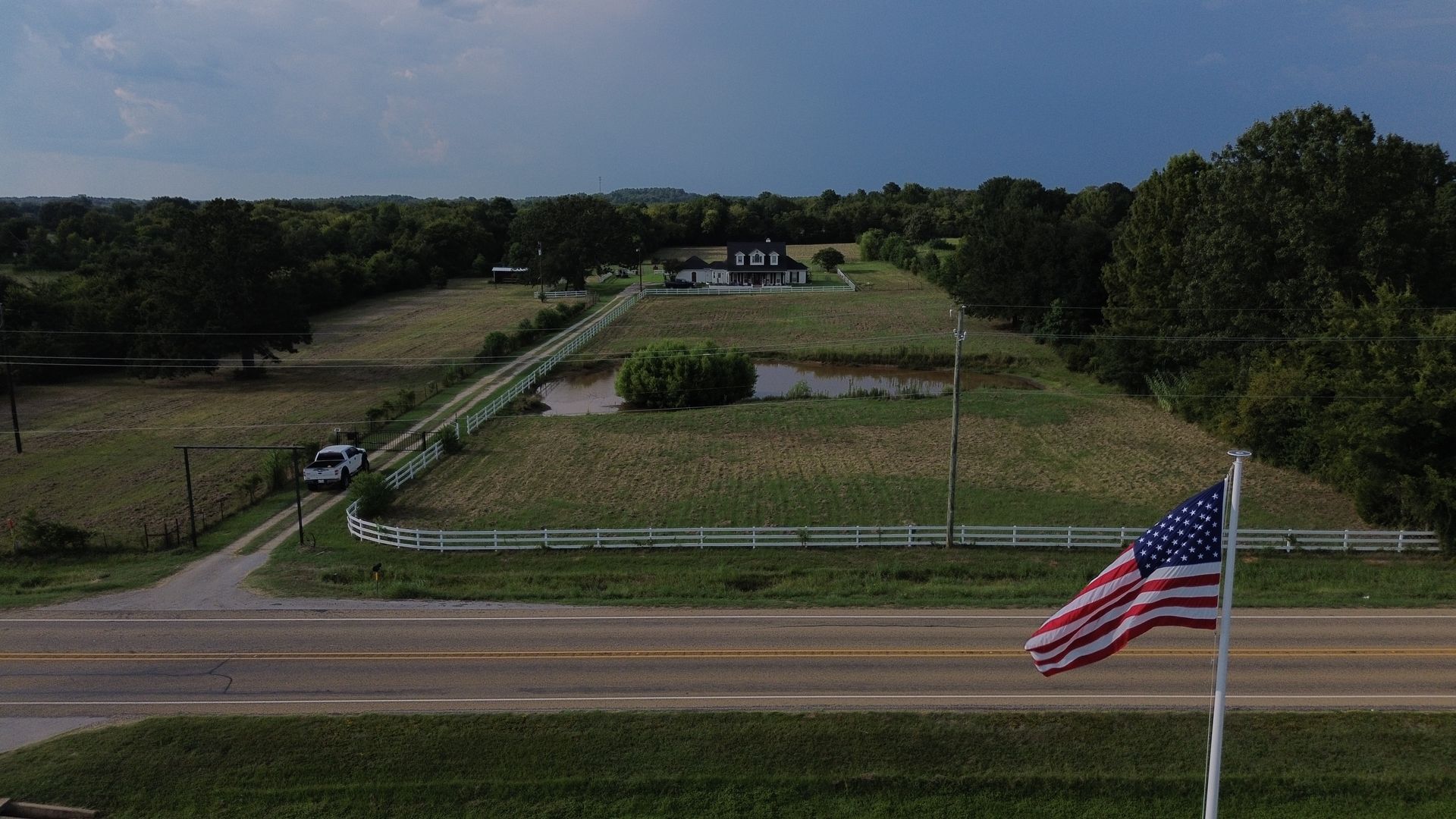 American flag waves in front of a rural property with a pond, road, and overcast sky.