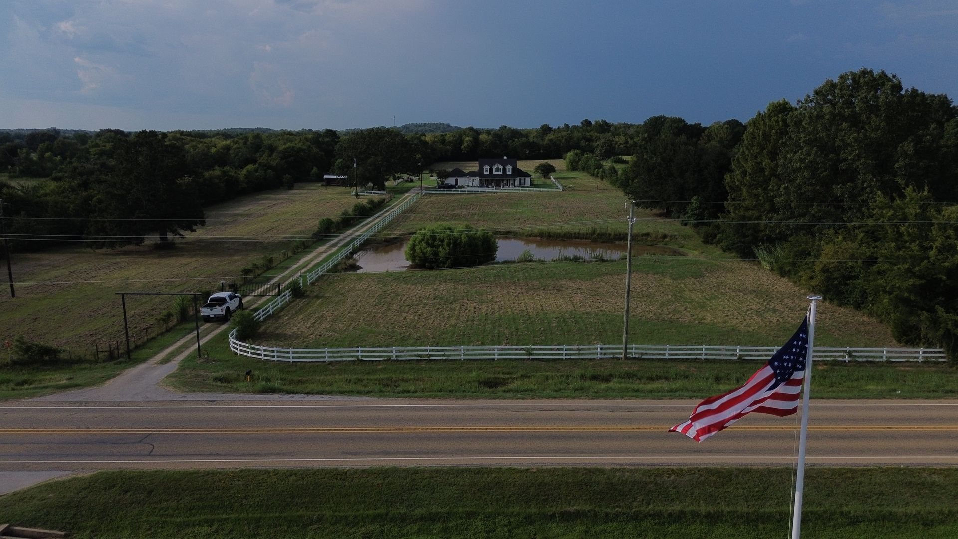 American flag waving, a house on a hill, open fields, and a dark cloudy sky in the background.